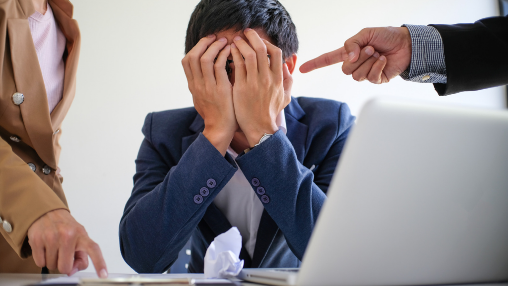 Workplace bullying isn't the problem of employees its management. The image shows a person under pressure with their head in their hands with managers pointing at them. 